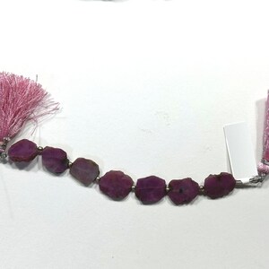 May include: A strand of raw, deep magenta-colored gemstone slices arranged in a row, separated by small metallic spacer beads. The strand is finished on both ends with light pink decorative tassels and includes a small white rectangular tag.