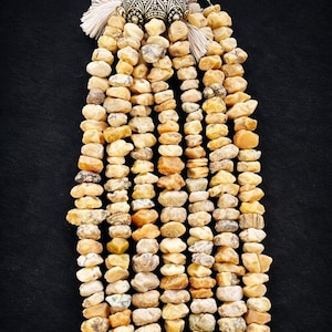 May include: A necklace composed of multiple strands of small, irregularly shaped, light-colored stones. The stones vary in shades of beige, tan, and light brown. The necklace is adorned with silver-toned metal accents and white tassels at the top.