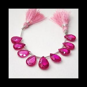 Op de afbeelding: Een ketting van helder roze, druppelvormige kralen met een facetgeslepen snit, geregen op een zilverkleurige draad. De ketting is geaccentueerd met twee roze kwastjes. De kralen zijn felroze.