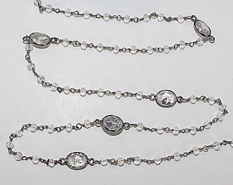 Könnte beinhalten: Eine lange Halskette mit einer dunklen Metallkette, die mit kleinen, klaren, facettierten Perlen und größeren, ovalen, klaren Steinen in dunklen Metallfassungen besetzt ist und ein wiederkehrendes Muster aus zarten Perlen und Akzenten bildet.