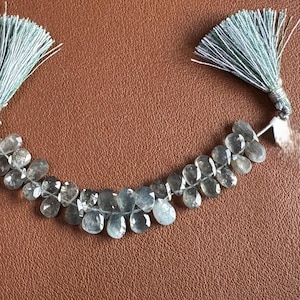 May include: A strand of light blue gemstone beads with a teardrop shape. The beads are strung together and are connected to two light blue tassels. The background is a brown leather-like surface.