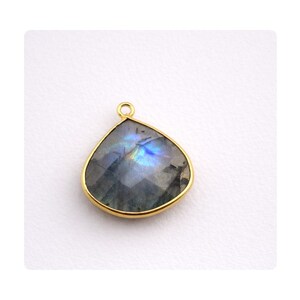 Pode incluir: Um pingente em forma de gota com uma moldura dourada. A pedra preciosa no interior tem uma mistura de cores cinza, azul e iridescente. O pingente tem um pequeno laço na parte superior para pendurar.