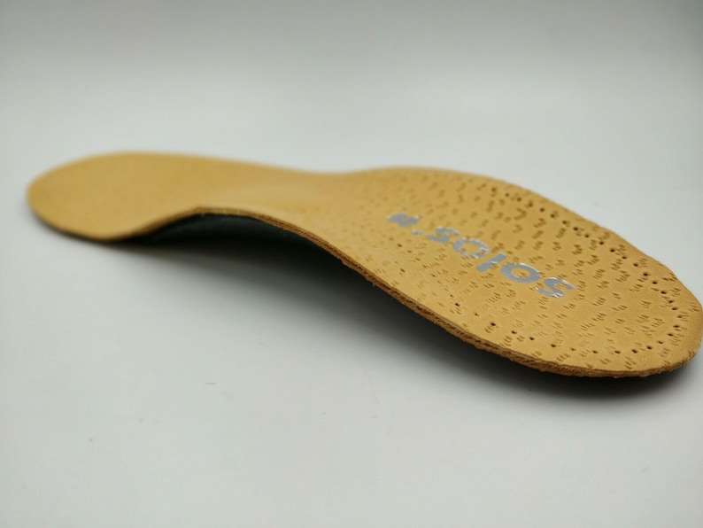 Op de afbeelding: Een beige leren inlegzool met een gestructureerd oppervlak en het woord "SOLOS" in zilver aan de zijkant.