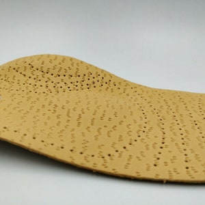 Op de afbeelding: Een beige leren inlegzool met een patroon van kleine gaatjes. De inlegzool is gebogen om de vorm van een voet te passen.