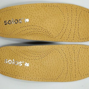 Op de afbeelding: Een paar beige inlegzolen met het woord "solos" in zilver op elke inlegzool.