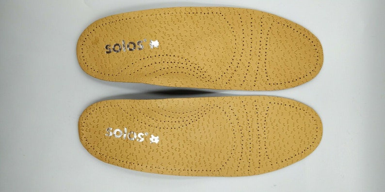 Op de afbeelding: Een paar beige inlegzolen met het woord "solos" in zilver op elke inlegzool.