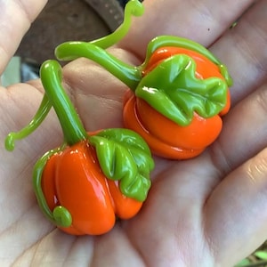 May include: Two small glass pumpkins with green stems and leaves. The pumpkins are orange and have a glossy finish.