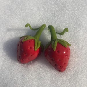 May include: Two red glass strawberries with green stems and leaves. The strawberries are sitting on a white surface.