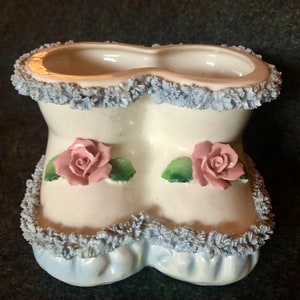 Könnte beinhalten: Ein weißer Keramik-Pflanztopf in Form von Babyschuhen mit blauem Besatz und rosa Rosen.