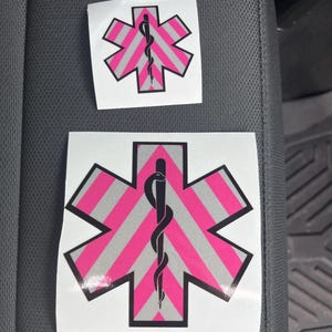 May include: Two pink and white reflective star of life stickers with a black snake and staff symbol in the center. The stickers are on a gray car seat.