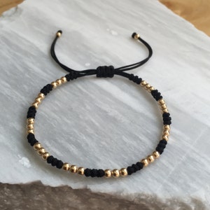 Può includere: Un braccialetto in corda nera con perline dorate. Il braccialetto è regolabile con un nodo scorrevole.