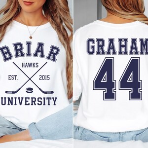 May include: A white t-shirt featuring a navy blue graphic on the front that reads BRIAR UNIVERSITY HAWKS EST. 2015 with crossed hockey sticks and a puck. The back of the shirt displays the name GRAHAM and the number 44 in large navy blue lettering.