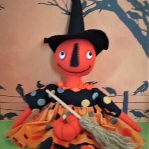 May include: A handmade Halloween doll with an orange head and body, wearing a black witch hat and a polka-dot dress. The doll holds a small pumpkin and a broom. The background features a Halloween-themed scene with crows.