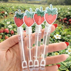 May include: Four clear bubble wands topped with strawberry-shaped stickers are held in a hand. The strawberry stickers have red fruit and green leaves. The background is a field of strawberries.