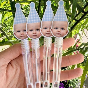 May include: Four bubble wands with baby face toppers wearing blue and white checkered party hats. The clear plastic tubes contain liquid and a wand for blowing bubbles. The background features green foliage and purple flowers.