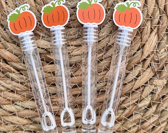 Op de afbeelding: Vier doorzichtige bellenblaasstokken met oranje pompoenopzetstukken zijn gerangschikt op een geweven mat. Roze lint krult rond de stokken. De pompoenopzetstukken hebben groene stelen en witte details.