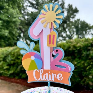 May include: A cake with white frosting and colorful sprinkles, topped with a decorative cake topper. The topper features a number 1, a sun, a popsicle, a flamingo, a beach ball, and the name "Claire" on a banner.