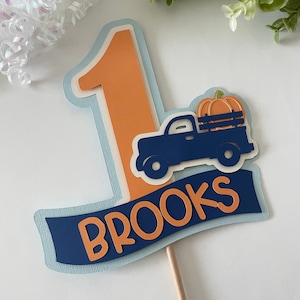 May include: A blue truck with an orange pumpkin in the bed, a blue banner with the name "BROOKS" in orange letters, and a large orange number "1" on a light blue background. This is a cake topper for a first birthday.