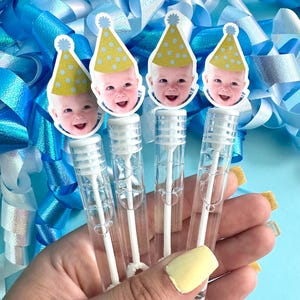 May include: Four clear bubble wands topped with baby faces wearing yellow party hats with blue polka dots. The wands are held in a hand with yellow painted nails, against a backdrop of blue and white ribbons.