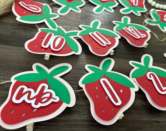 May include: A string of red and green strawberry-shaped paper cutouts with white numbers 1 through 12. The strawberries are strung together with twine and have wooden clothespins attached to them.