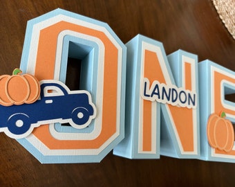 May include: A blue, orange, and white papercraft "ONE" with a truck carrying a pumpkin and the name "LANDON" on the "N".