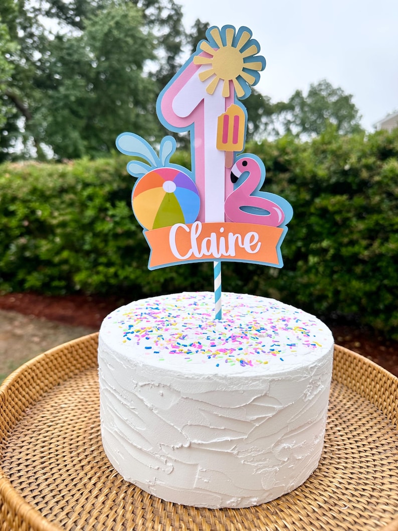 May include: A white frosted cake with colorful sprinkles, topped with a custom cake topper. The topper features a number one, a sun, a flamingo, a beach ball, and the name "Claire" in orange. The cake is on a woven tray.
