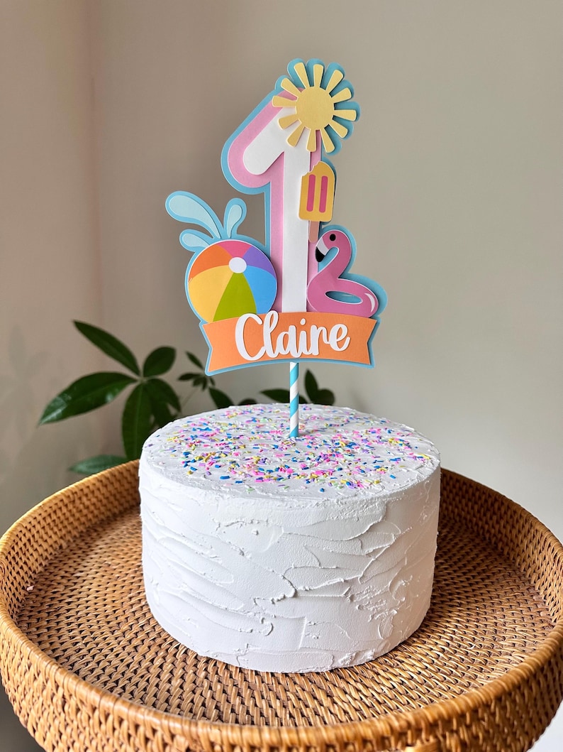 May include: A white frosted cake with colorful sprinkles, topped with a decorative cake topper. The topper features the number 1, a sun, a flamingo, a beach ball, and the name "Claire" on an orange banner. The cake sits on a woven tray.