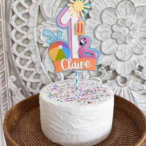 May include: A white frosted cake with colorful sprinkles, topped with a decorative cake topper. The topper features a number one, a sun, a flamingo, a beach ball, and the name "Claire" on an orange banner. The cake sits on a woven tray.