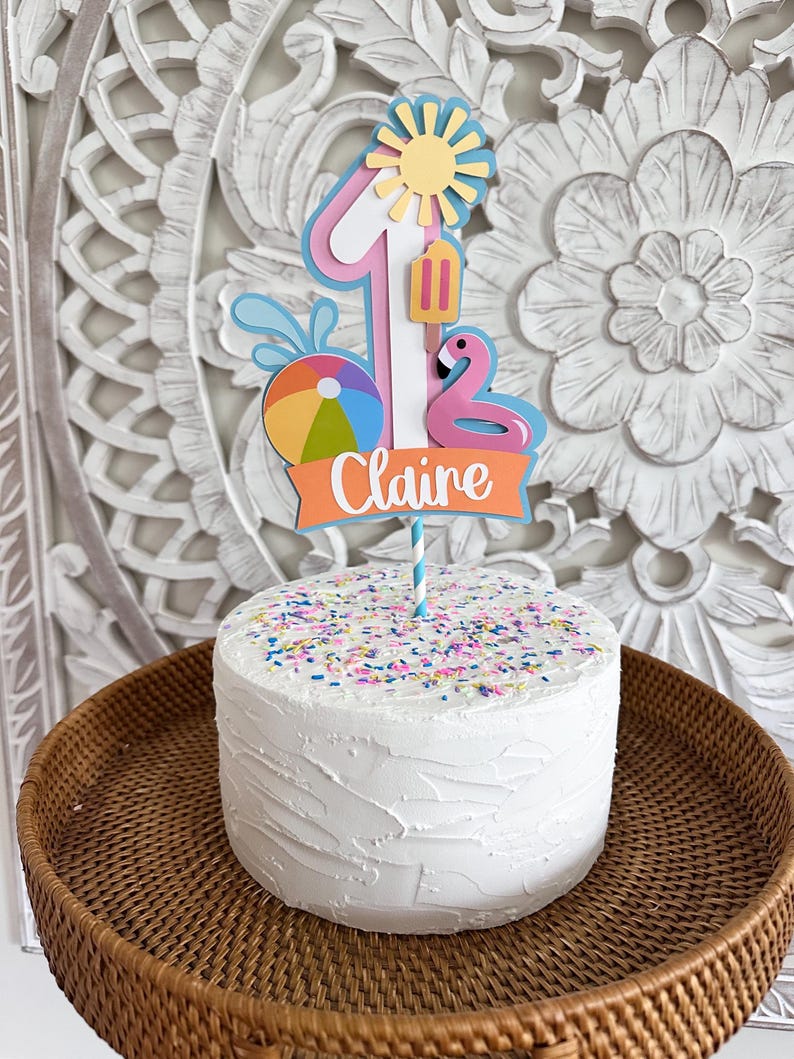 May include: A white frosted cake with colorful sprinkles, topped with a decorative cake topper. The topper features a number one, a sun, a flamingo, a beach ball, and the name "Claire" on an orange banner. The cake sits on a woven tray.