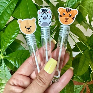 May include: Three clear bubble wands with animal-themed toppers: a bear, a zebra, and a tiger. The wands are held against a backdrop of green leaves. The bear is tan, the zebra is gray and white, and the tiger is orange and brown.