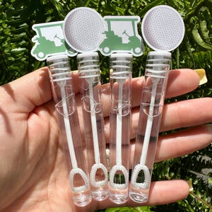 May include: Four clear bubble wands with white wands and bubble wands. Each wand has a golf ball and golf cart design on top. The wands are held in a hand against a background of green leaves.