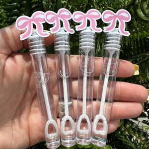 May include: Four clear bubble wands with white wands and bubble loops. Each wand is topped with a pink bow cutout. The wands are held in front of a green leafy background.