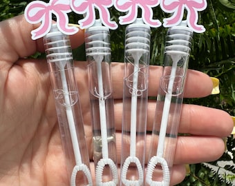 May include: Four clear bubble wands with white wands and bubble loops. Each wand is topped with a pink bow cutout. The wands are held in front of a green leafy background.