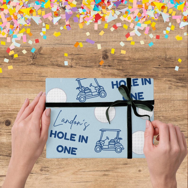 Op de afbeelding: Een lichtblauwe geschenkdoos met een zwart lint en strik, met illustraties van golfkarretjes en de tekst "Landon's Hole in One". De doos is versierd met witte golfbalontwerpen. Bunte confetti is erboven verspreid.