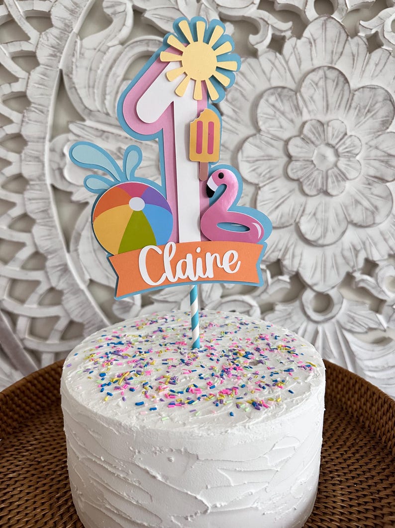 May include: A white frosted cake with colorful sprinkles, topped with a decorative "1" cake topper. The topper features a sun, popsicle, flamingo, beach ball, and the name "Claire" on an orange banner. The cake is on a woven tray.