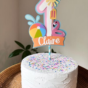 May include: A white frosted cake with colorful sprinkles, topped with a decorative "1" cake topper. The topper features a sun, popsicle, flamingo, beach ball, and the name "Claire" on an orange banner. The cake sits on a woven tray.
