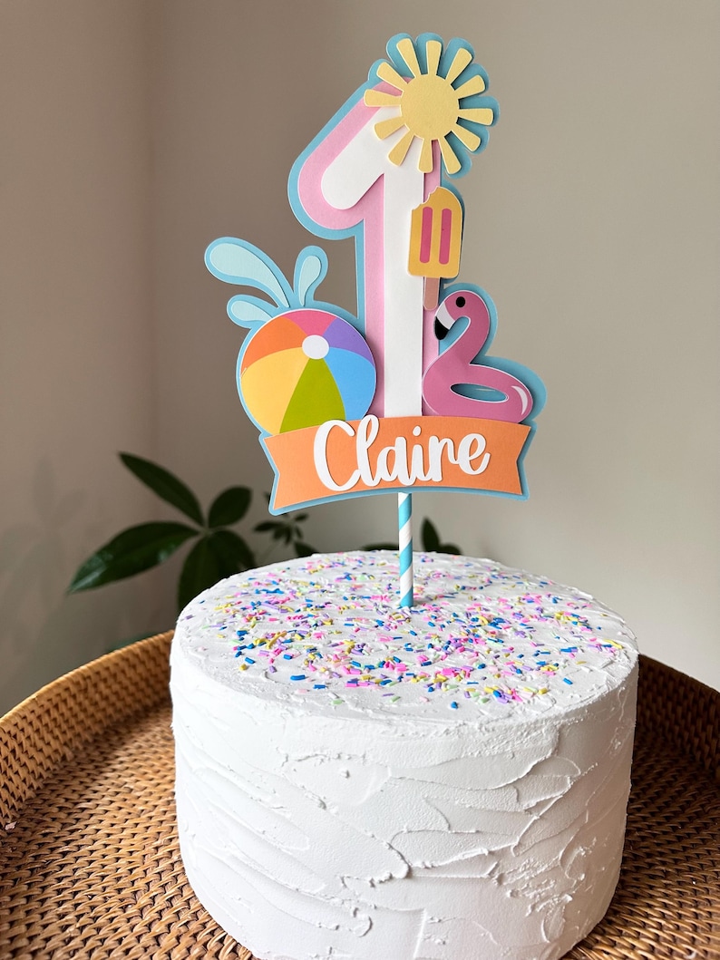 May include: A white frosted cake with colorful sprinkles, topped with a decorative "1" cake topper. The topper features a sun, popsicle, flamingo, beach ball, and the name "Claire" on an orange banner. The cake sits on a woven tray.