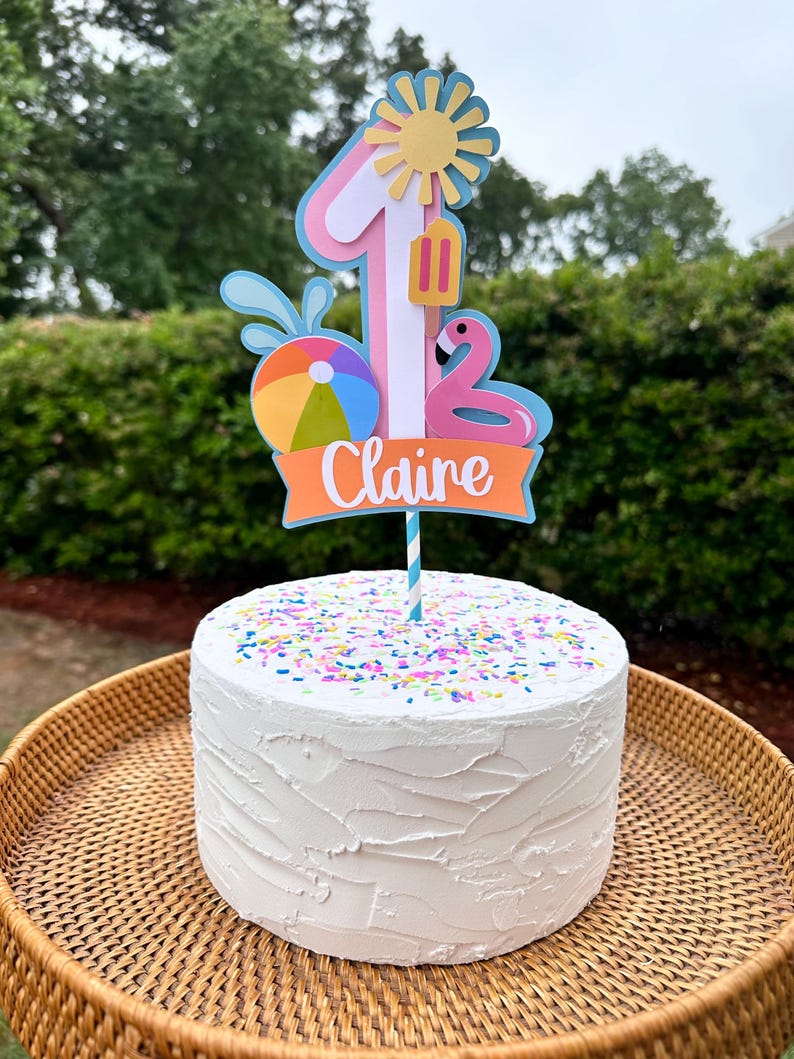 May include: A white frosted cake with colorful sprinkles, topped with a decorative cake topper. The topper features the number 1, a sun, a flamingo, a beach ball, and the name "Claire" in white lettering on an orange banner. The cake sits on a woven tray.