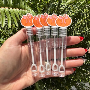 May include: A hand holds five clear bubble wands, each topped with a pumpkin sticker featuring an orange pumpkin and a pink bow. The wands have white bubble wands and are set against a backdrop of green foliage.