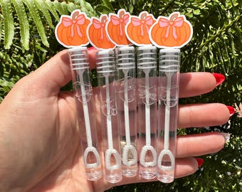 Puede incluir: Cinco varitas de burbujas transparentes, cada una con una pegatina de calabaza con una calabaza naranja y un lazo rosa. Las varitas tienen varitas de burbujas blancas. Sobre un fondo de follaje verde.
