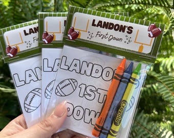 May include: Clear plastic bags containing coloring pages and crayons. Each bag has a label with the text "LANDON'S First Down" and football graphics. The crayons are orange, blue, and green.
