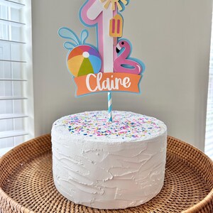 May include: A white frosted cake with colorful sprinkles sits on a woven tray. A decorative cake topper features a number 1, a sun, a popsicle, a flamingo, and a beach ball. The name "Claire" is written on the topper.