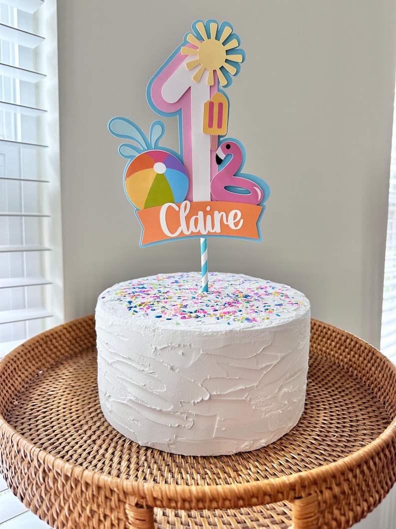 May include: A white frosted cake with colorful sprinkles sits on a woven tray. A decorative cake topper features a number 1, a sun, a popsicle, a flamingo, and a beach ball. The name "Claire" is written on the topper.