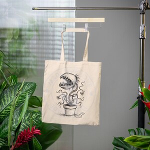 May include: A beige tote bag with a black and white illustration of a carnivorous plant with a toothy mouth. The plant is in a pot and has a sock on the ground next to it.