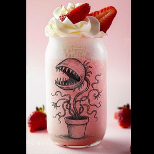 Op de afbeelding: Een roze aardbeien milkshake met slagroom en aardbeien erop. Het glas heeft een zwart-witte illustratie van een vleesetende plant met een grote mond.