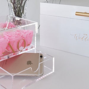 May include: A white box with the words "Our Wedding" written in gold script. The box is sitting on a table with a clear acrylic box containing pink roses and a gold XO. A clear acrylic box with a mobile phone is sitting on top of the box with the roses.