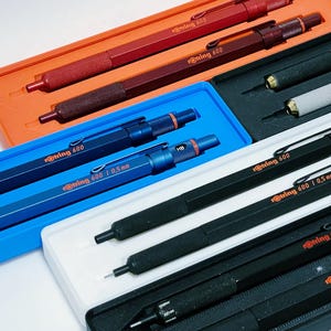 May include: A collection of Rotring 600 mechanical pencils in various colors, including red, blue, and black, displayed in their original packaging. The pencils have a distinctive hexagonal barrel design and are labeled with their respective sizes.