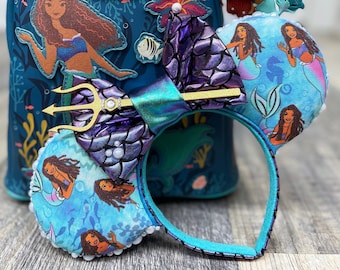 May include: A pair of blue and purple Disney Loungefly Minnie Mouse ears with a mermaid theme. The ears feature a gold trident, purple and blue scales, and images of Ariel and other mermaids. The ears are attached to a blue headband.