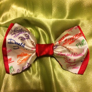May include: A white satin bow tie with red trim, featuring colorful handwritten messages from Disney characters, including Donald Duck, Mickey Mouse, and Pluto. The bow tie has a red satin center.