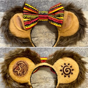 May include: Lion-themed ear headband with faux fur, tan ears, and a patterned bow. The headband features a brown faux fur mane, tan inner ears, and a bow with red, yellow, and black tribal patterns. The second headband has a lion cub and sun symbol.